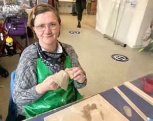 Louise is sat a table wearing a green apron. She is holding some clay she is working on and smiling. 