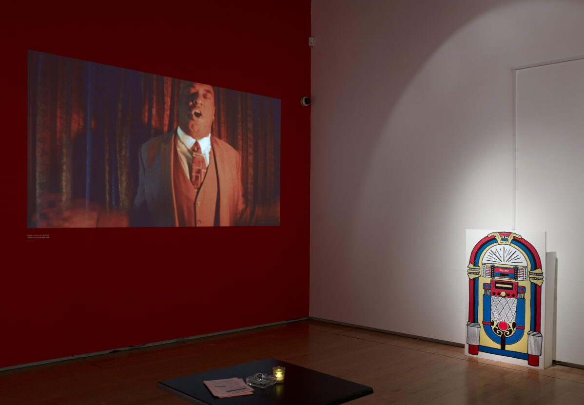 Projected film screen on a gallery wall, showing a man singing, alongside a cardboard juke box.