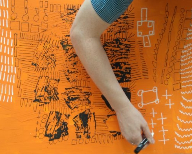 An arm holding a white pen leaning over a large piece of art painted orange.