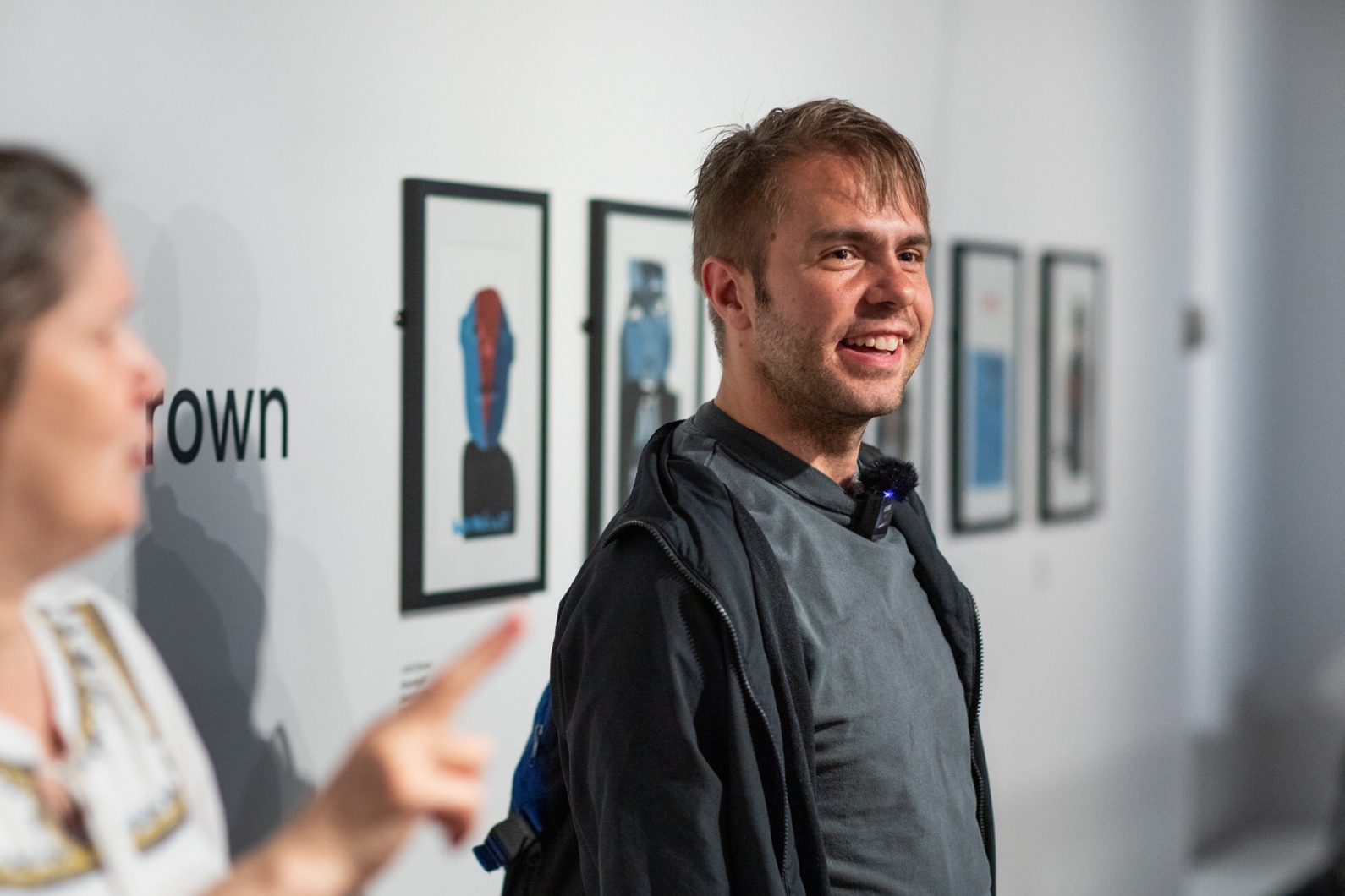 Young man, artist Liam Ashworth, stood in a gallery in front of the framed pictures in the wall. He is wearing a microphone and is smiling.