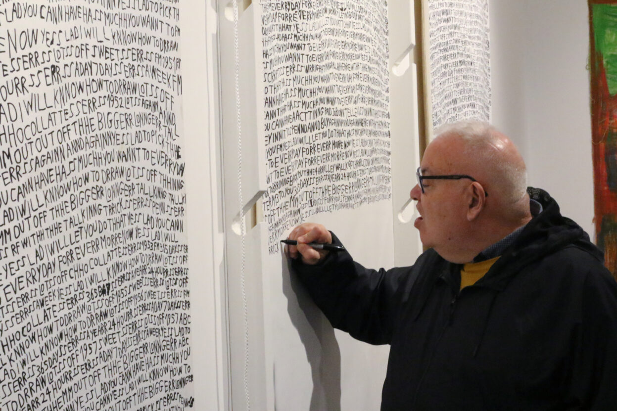 Barry Anthony Finan working during the 'YESS LAD' exhibition launch - he is writing with black pen on paper attached to the wall