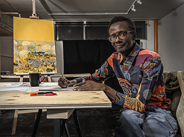 Leslie Thompson sitting at a desk drawing, with one of his artworks displayed on an easel