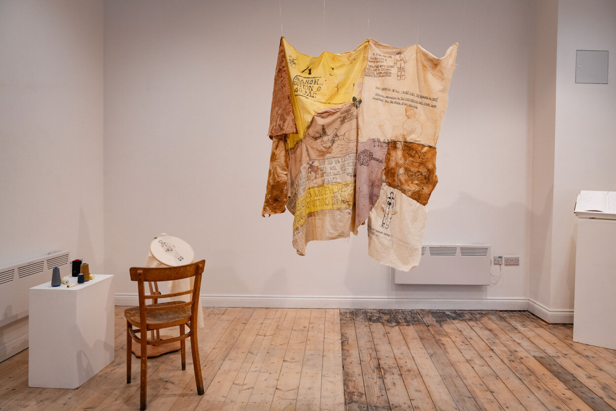 A piece of textile art by Sarah Lee. It is hanging up in the centre of a room. There is also a wooden chair and a small embroidery work at the side.