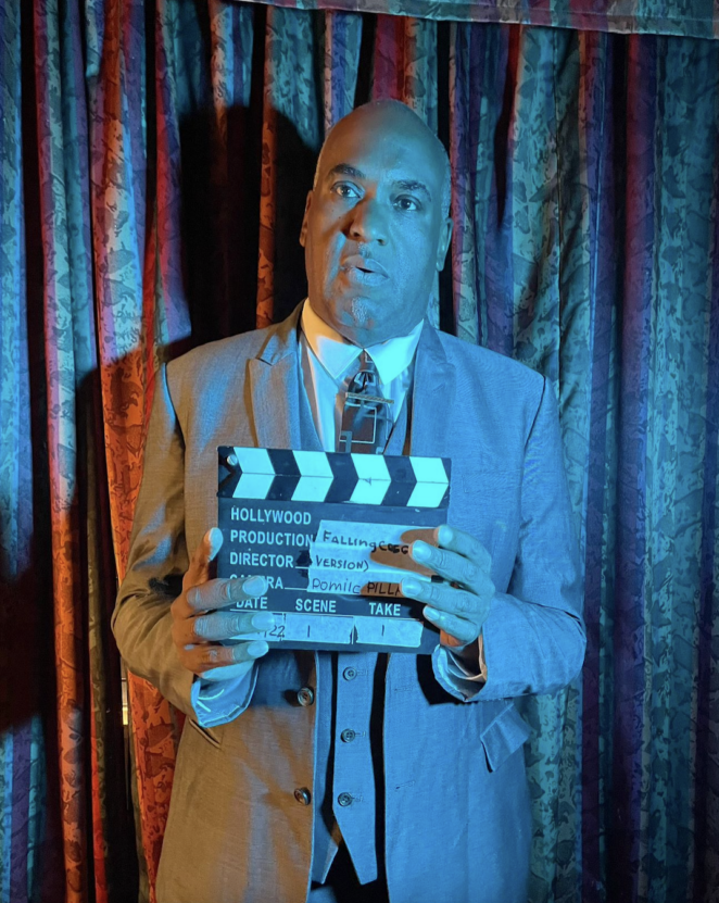 Man in suit standing under a spotlight in front of patterned curtains holding a clapper board