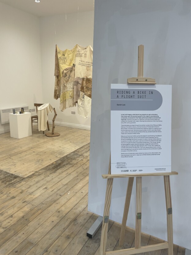 Sarah Lee's hand embroidered textile artwork is hung up in the middle of a studio space alongside a small embroidery artwork. There is a wooden easel with a description of the artwork too.