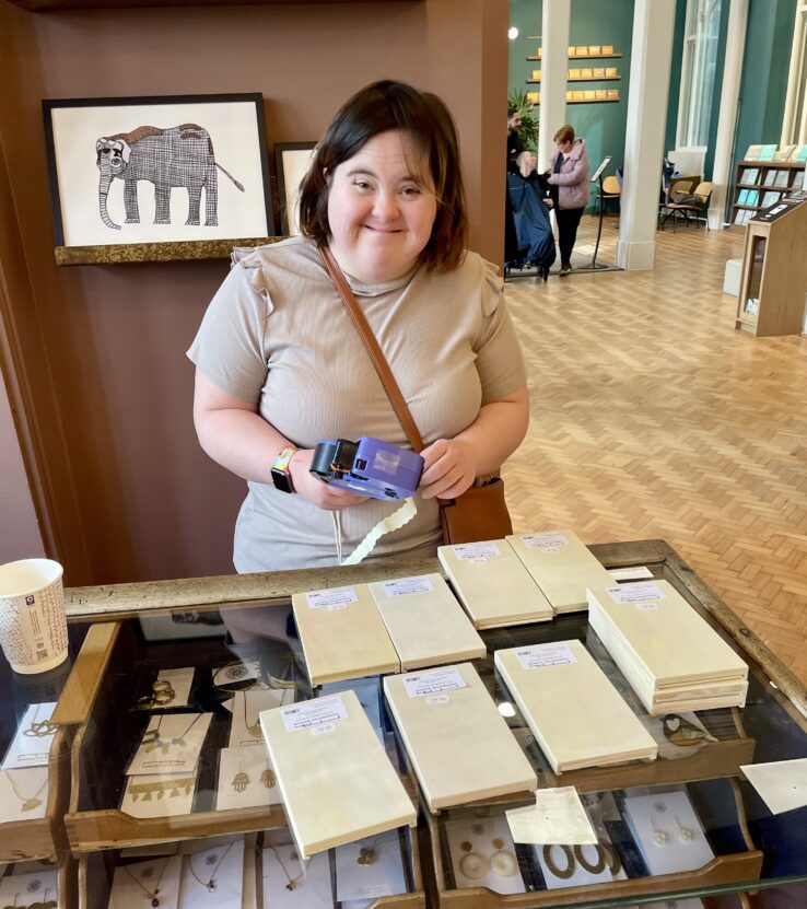 Artist Amy Ellison stood behind a desk at the Manchester Museum Shop pricing items. She is smiling at the camera.