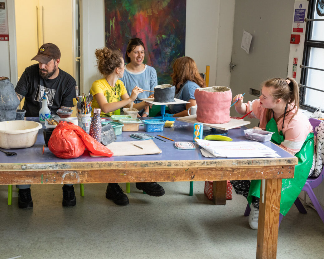A group of adults and young people sat round a large table of art materials, with two girls making clay pots.