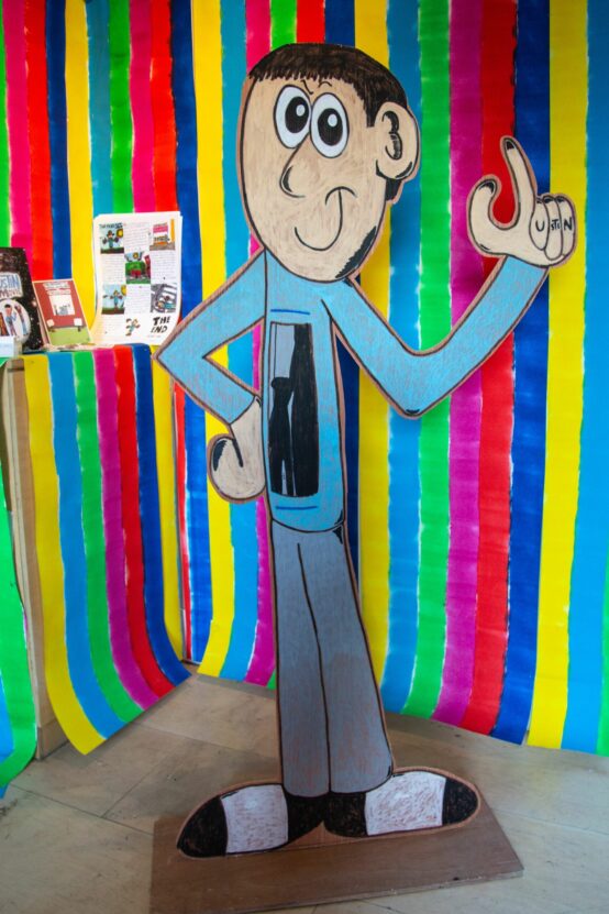 Freestanding cardboard cut out of a cartoon man, in front of bright vertical stripes, at the Behold Room exhibition