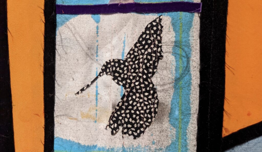 A textile artwork featuring a black and white bird on a blue, white and orange background