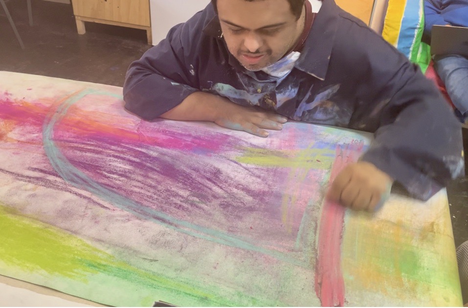 Artist sat at a table in front of a large piece of pastel artwork.