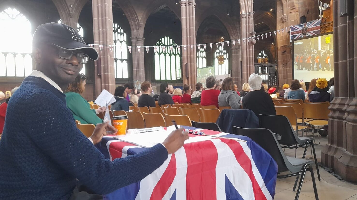 A man, Leslie Thompson, sat drawing at a table with a union jack flag on it in Manchester Cathedral.