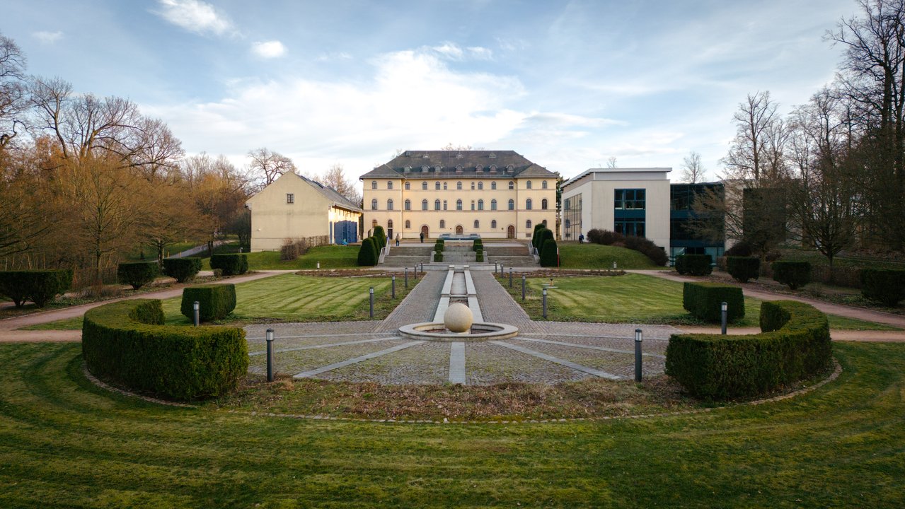 A large cream coloured building with many windows, with a lawn and long drive in front of it.