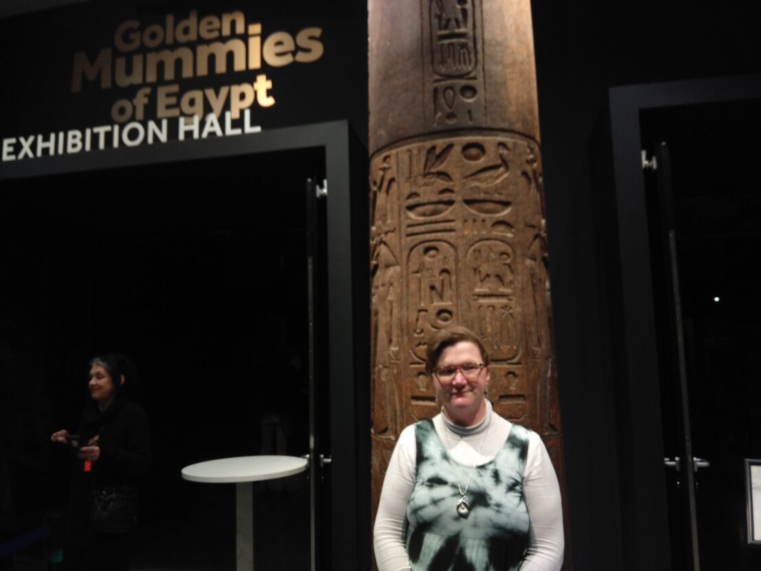 Louise Hewitt stood at the entrance to the Golden Mummies exhibition, stood in front of a pillar inscribed with hyroglyphs.
