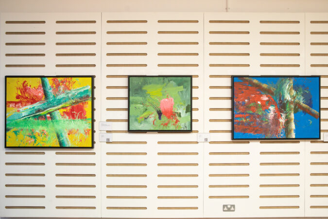 Three pieces of artwork hung on a wall at an exhibition. Abstract paintings, one with yellow background and red and green paint on top. One mainly green with a mix of green paint. One mainly blue with red and brown paint.