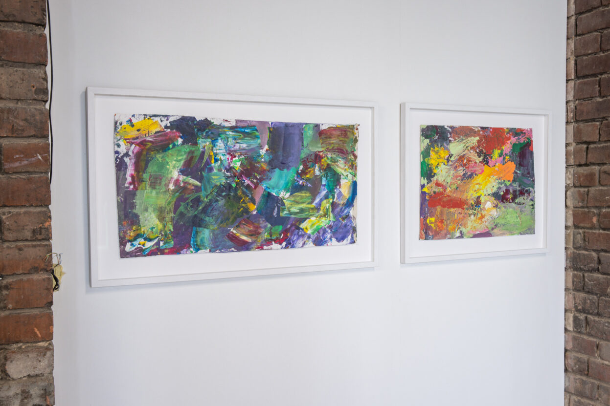 Two abstract paintings in white frames on a white gallery wall.
