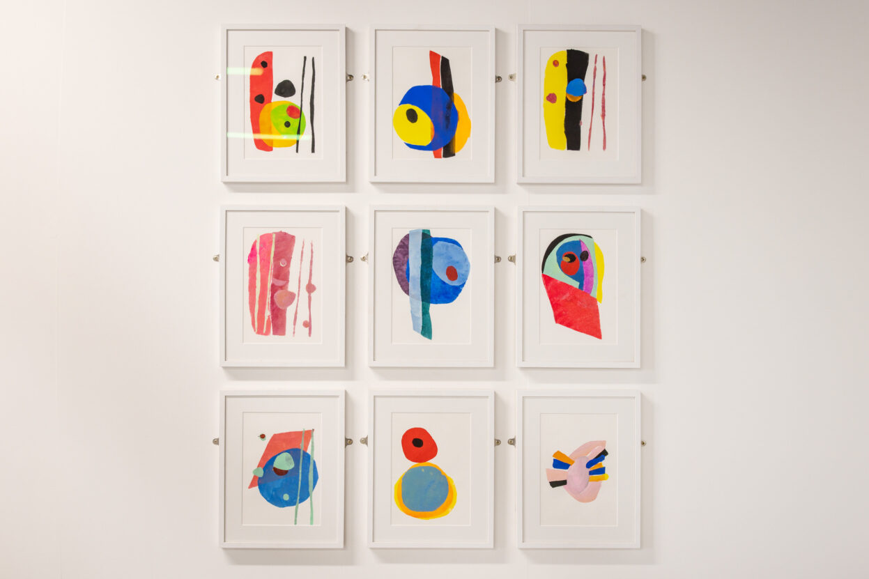 A collection of 9 colourful abstract stencil prints in white frames hung on a white gallery wall.