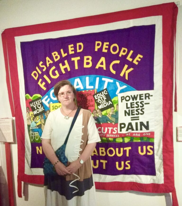Woman stood in front of a banner saying 'disabled people fightback'.