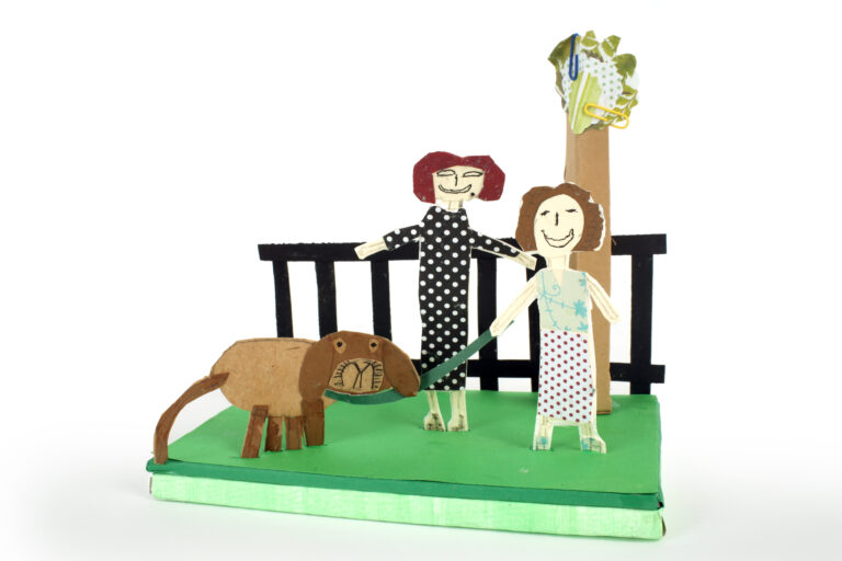 A 3D paper sculpture of two women waling a doh with a tree and a fence.