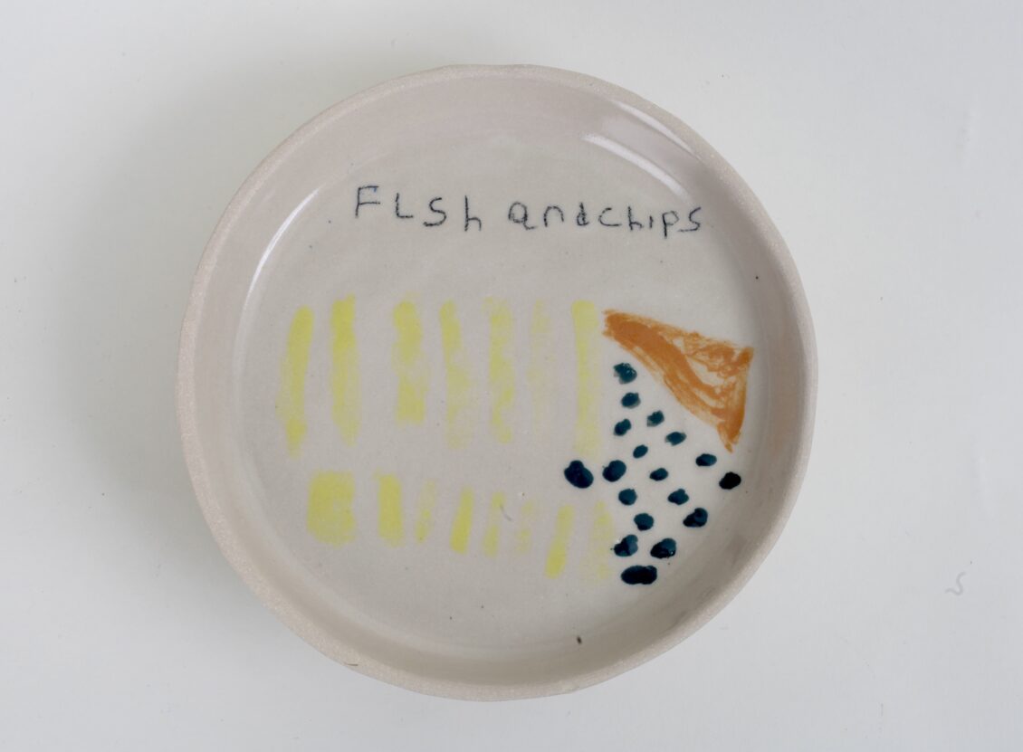 A round white ceramic plate painting with a picture of fish and chips and words saying 'fish and chips'.