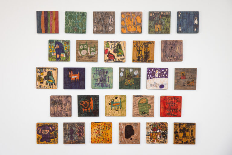 A collection of small wooden square panels hung on a white gallery wall, with illustrations of various different characters.