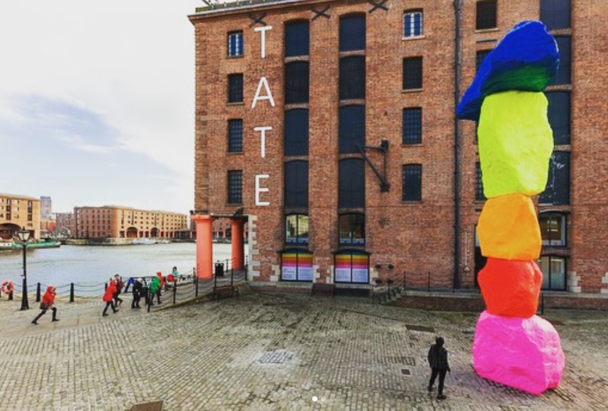 The exterior of Tate Liverpool, a large brick building with the words 'tate' written on the outside and a large colourful sculpture in the foreground.