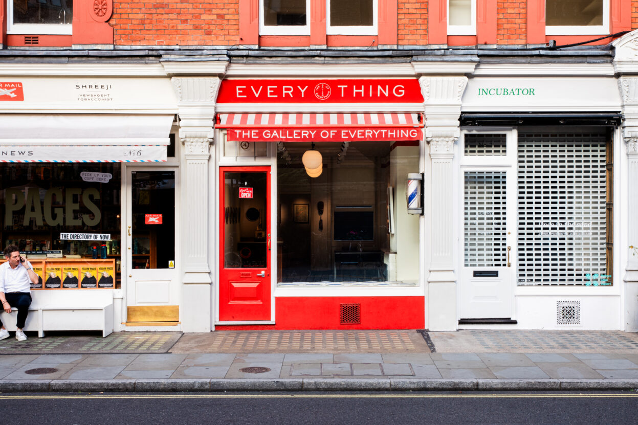 The shop front of an art gallery with a large window and red door.