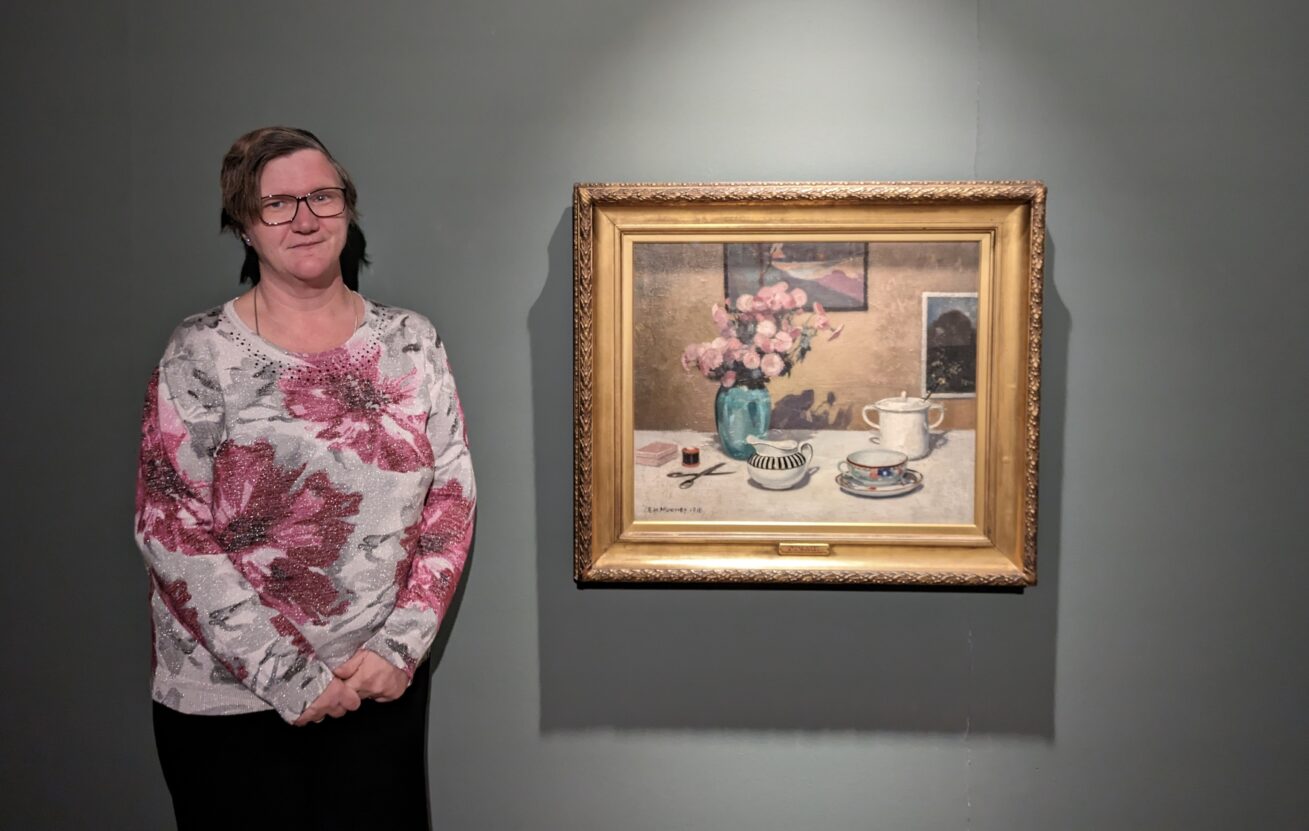 Artist Louise Hewitt stood in front of a framed picture of flowers on a gallery wall
