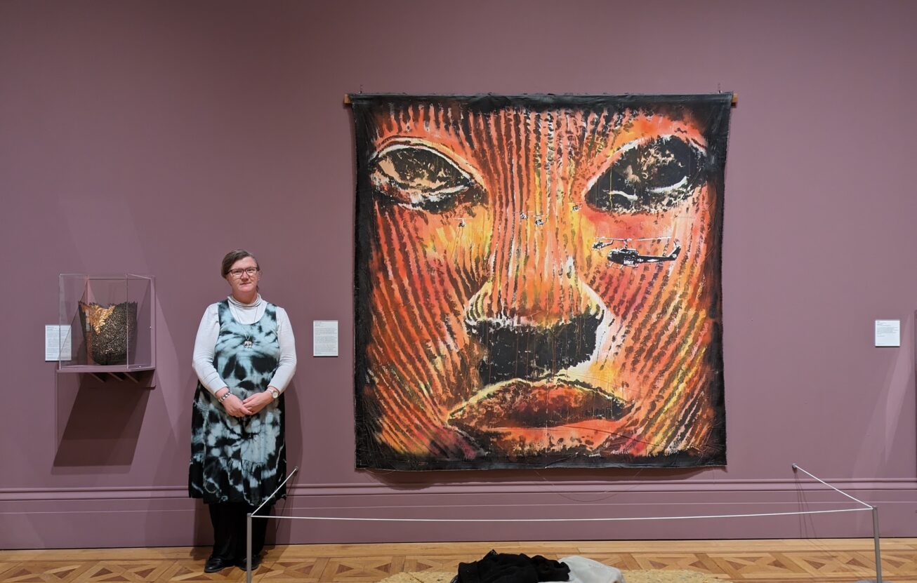 Artist Louise Hewitt stood next to a large painting of an african mask on a purple gallery wall.