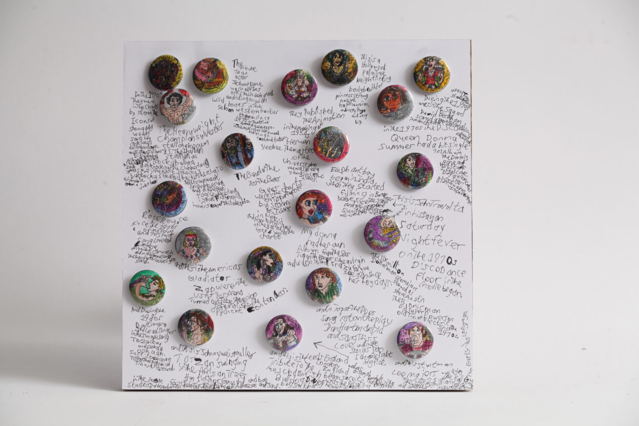 A selection of small hand drawn pin badges presented on a white board, with the artist's writing describing each badge.