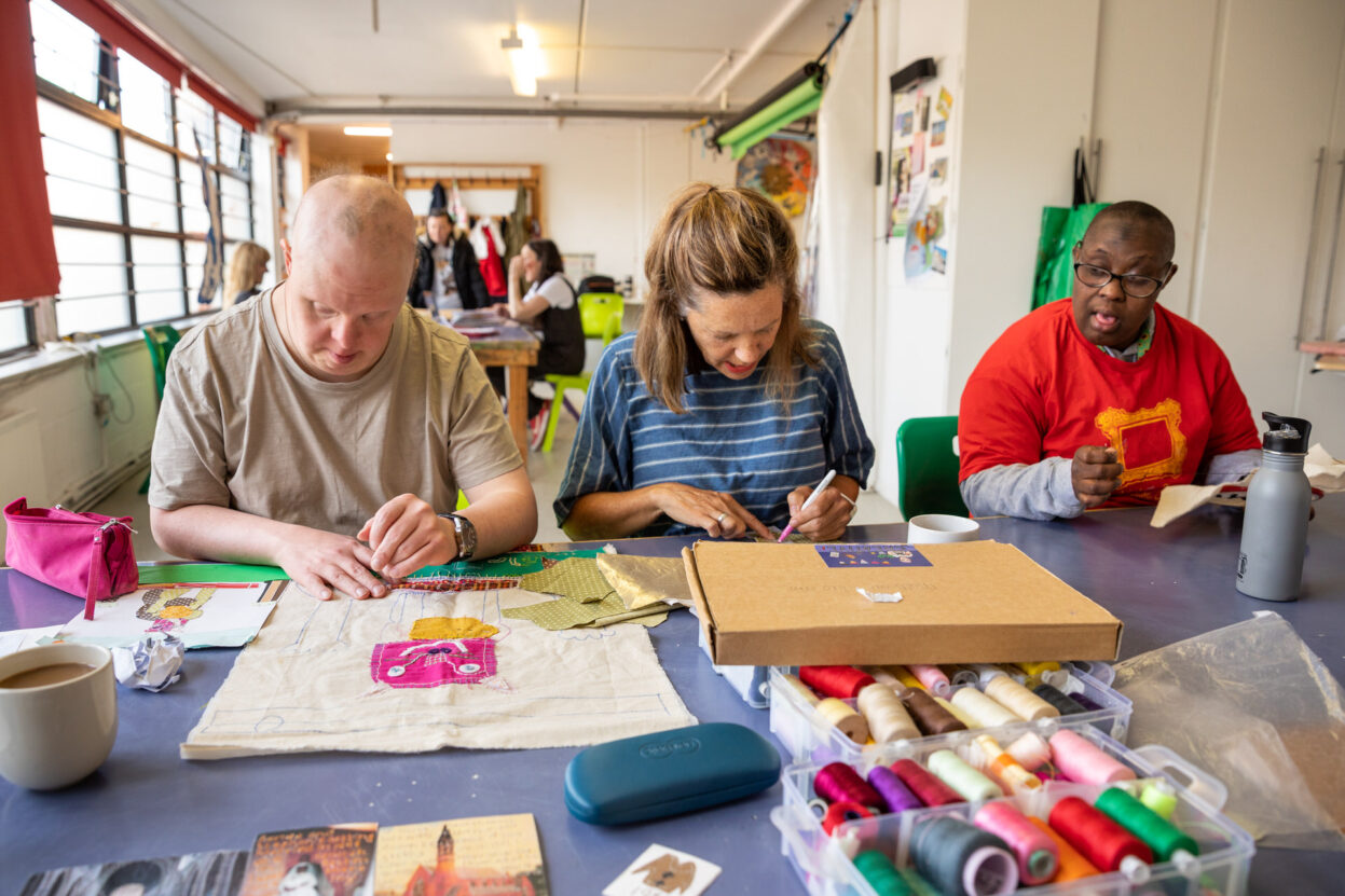 Three people sat at a table working on textile art projects.