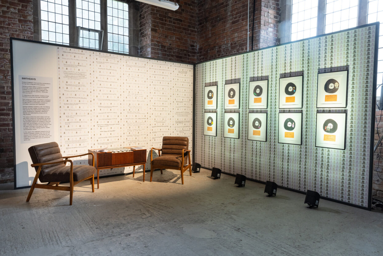 A section of the We Are Gathered Here Together exhibition with two chairs, a record player and artwork on the walls.