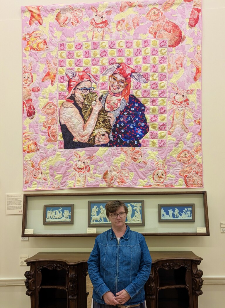 Louise Hewitt stood in front of a large pink textile artwork of two women, one of which is holding a rabbit.