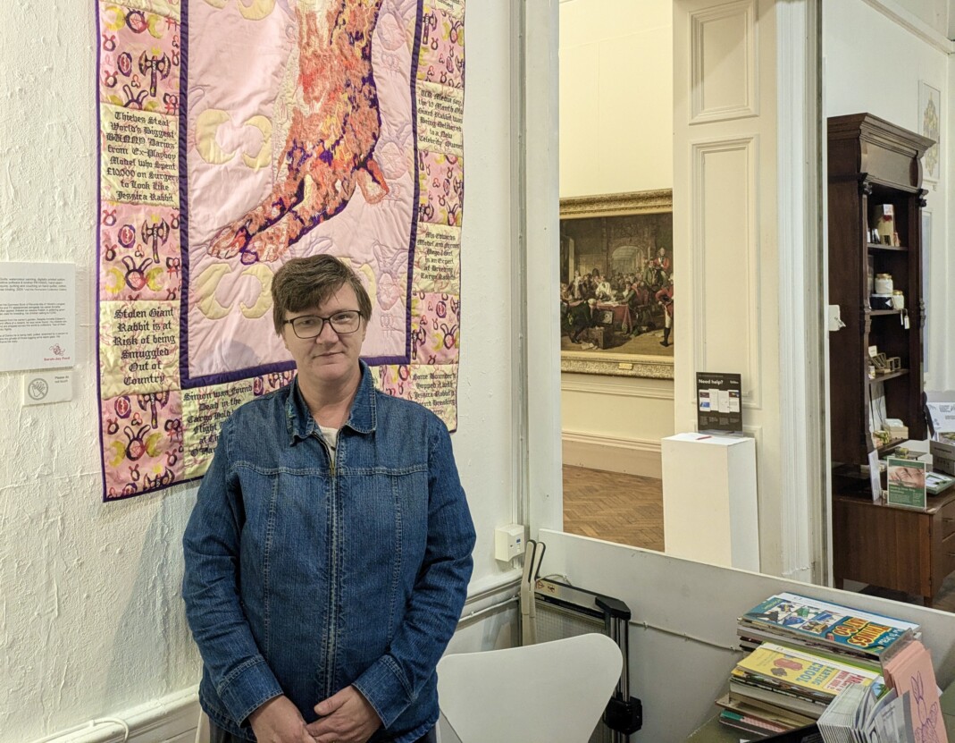 Louise Hewitt stood in from in front of a pink textile hanging, hung on a white gallery wall.