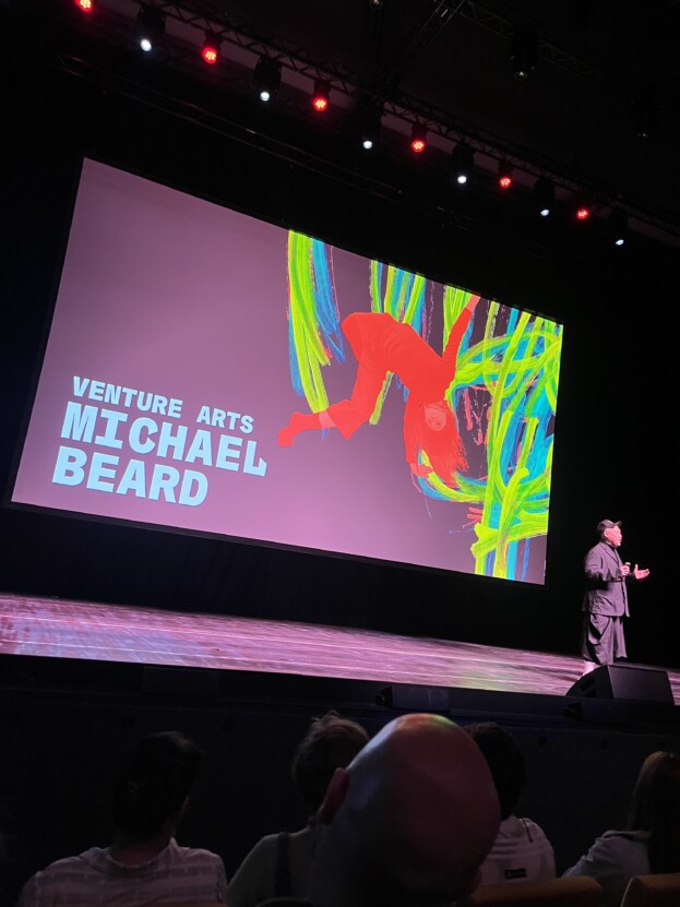 Large screen on a stage promoting the Michael Beard exhibition with a man stood on the stage.