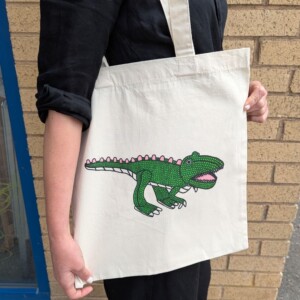 A woman holding a white tote bag with a dinosaur illustration by Andrew Johnstone