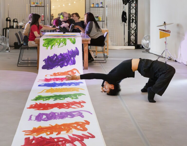 A long roll of paper with different paint on it and a dancer leaning back towards the paper. The artist - Michael Beard - sits at a table painting.