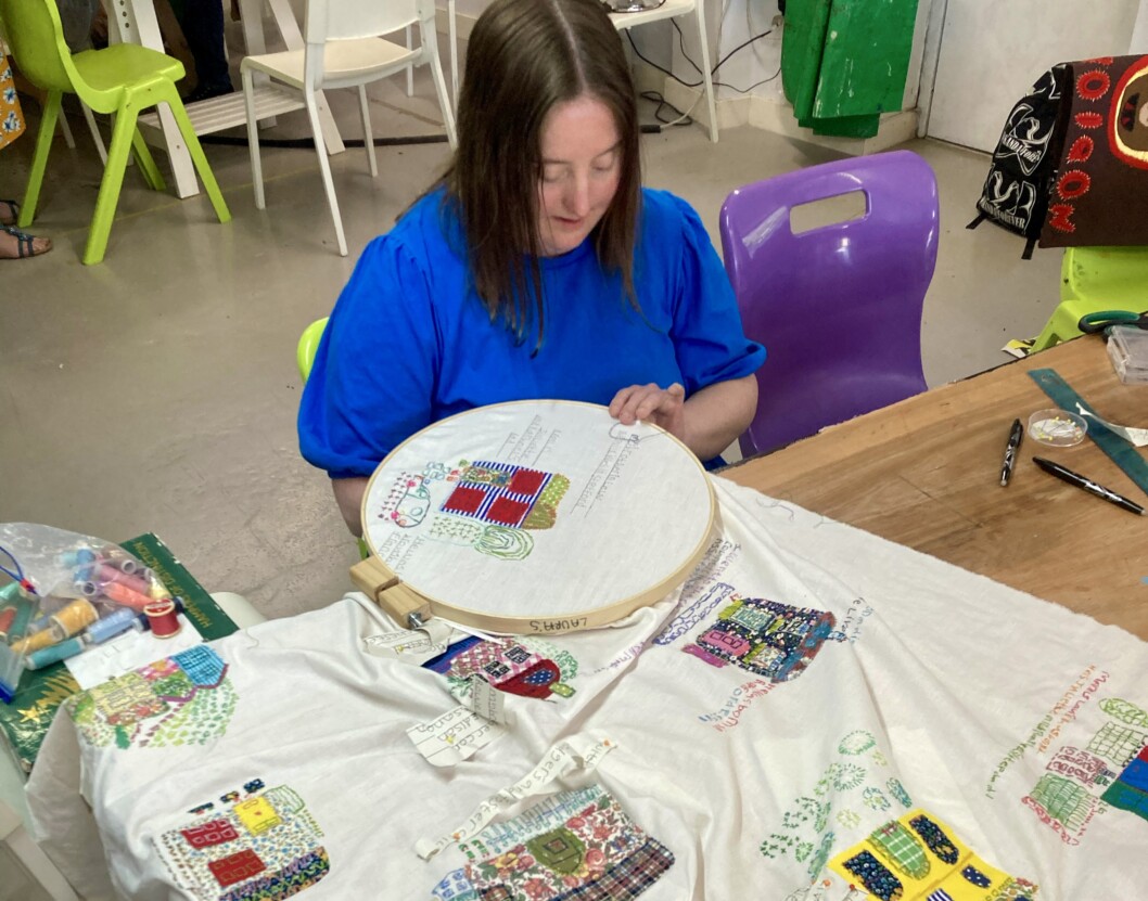 A young woman with brown hair and wearing a blue tshirt sat in front of a table sewing.