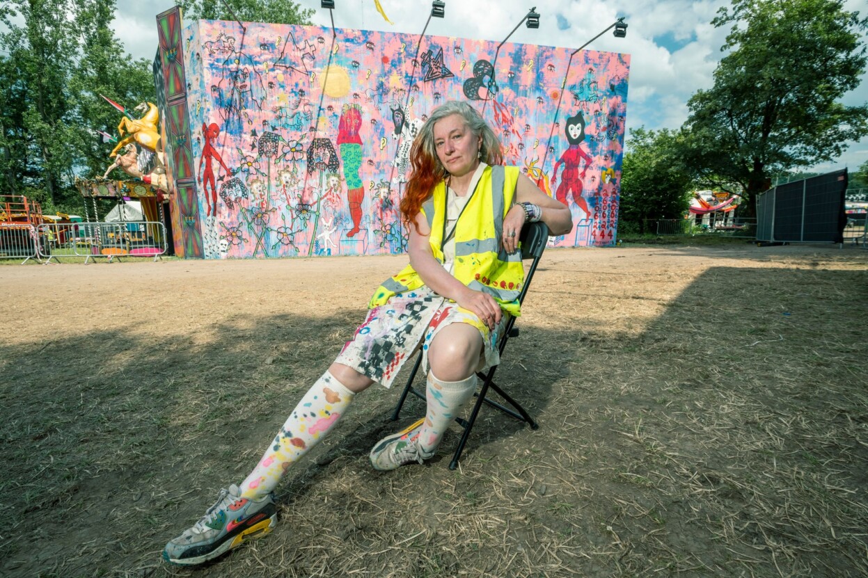The artist Delaine Le Bas sat on a chair on a piece of grass outside at Glastonbury Festival. There is a large-scale brightly coloured mural on the wall behind them.