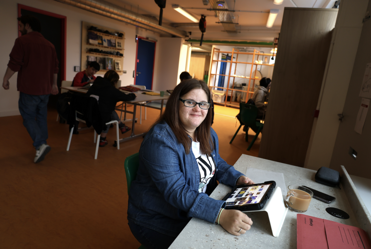 Venture Arts studio artist Nevaeh sits at a table with an eye pad smiling. She wears a white t-shirt with a blue denim shirt over the top, and she is wearing glasses.