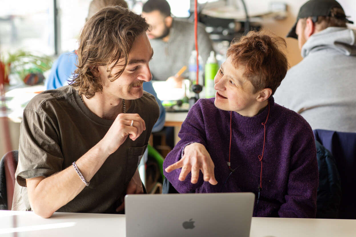 Two people are sat at a table talking and looking at a laptop.