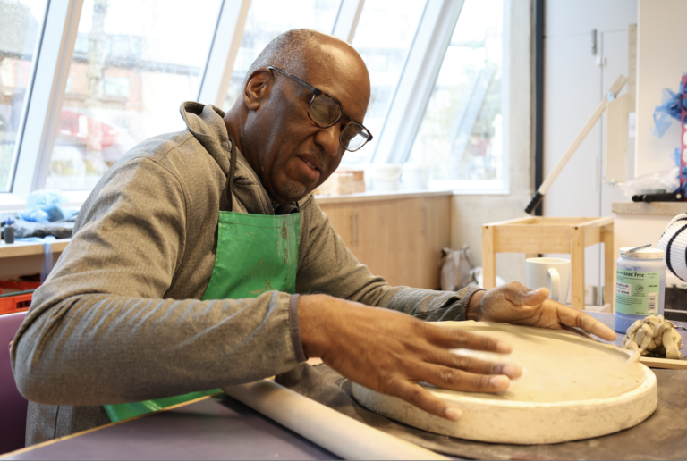 An artist sits at a table with a green apron on making a ceramic plate in the Venture Arts studio.