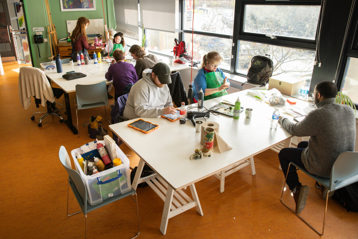 Studio 4 at Venture Arts. There are two white tables with artists sat at them working. There are large windows running along the wall of the studio.