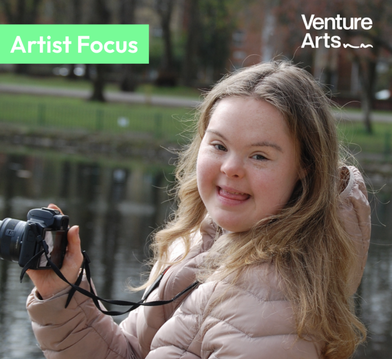 Venture Arts studio artist Maddi Lingard wearing a coat, holding a camera and smiling. The Venture Arts logo is the the top right corner and the words "Artist Focus" are in the top left corner.